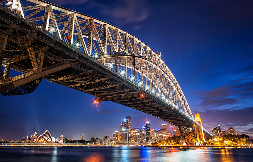 The Sydney Harbour Bridge viewed from underneath, the North Sydney side of the harbour, at night. The Opera House and city lights shine on the other side of the harbour.