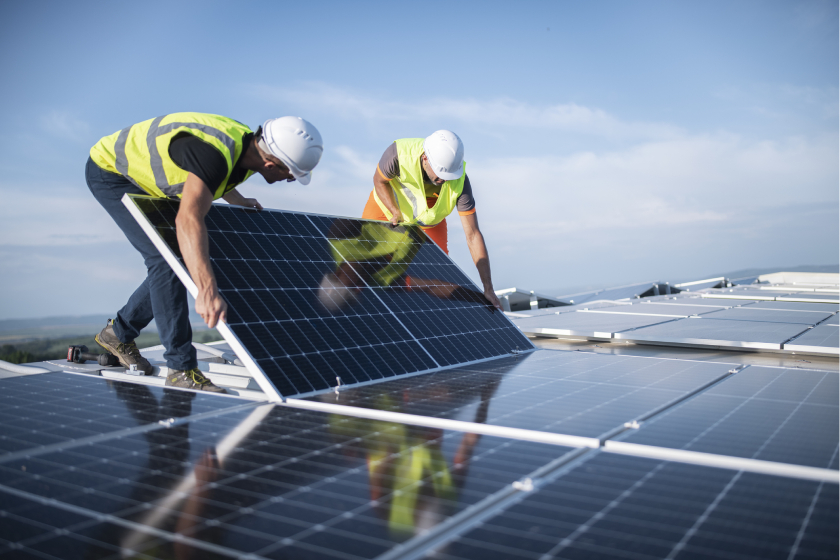 construction workers installing a solar panel. The mood of the photo is focused. They are both handling the panel with extreme care and precision. They are wearing bright construction vests and safety gear. 