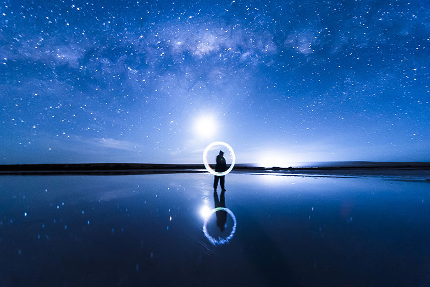 A person stands on a salt lake against a dark background, under a full moon and the milky way, and paints a glowing circle.