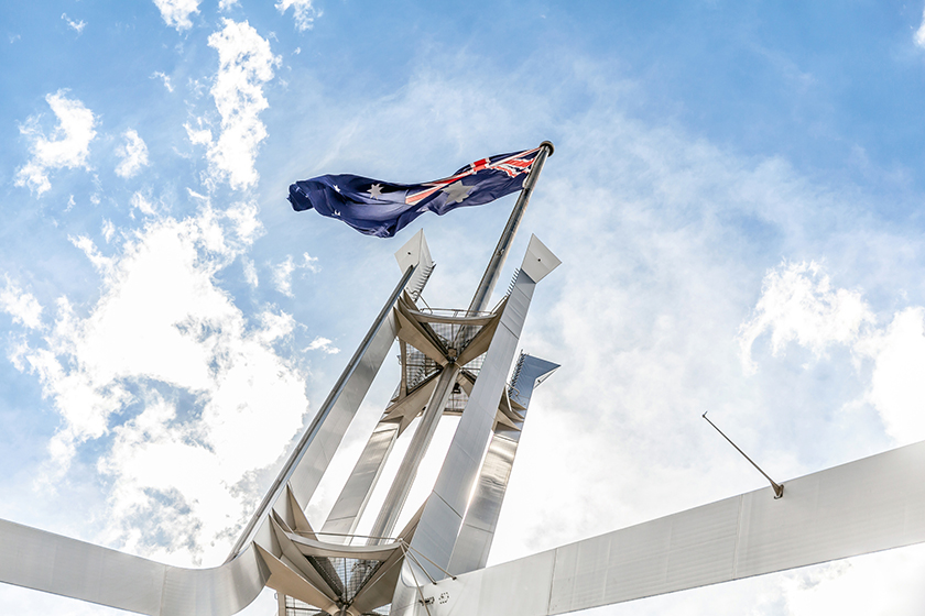The Australian flag atop Parliament House, Canberra, shot from below with a bright blue clear sky behind.