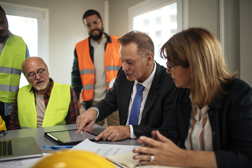 In a construction site office, accountant shows construction business owner and staff information on a tablet device.