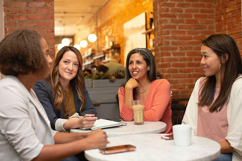 A group of four professional women sit around a table, speaking collaboratively and looking happy.